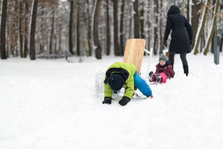 Photo for Joyful Little Brother and Sister in Warm Clothes Happily Playing With Mother and Falling Down From Sledding - Royalty Free Image