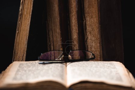 Photo for An open blurred page with hebrew religious text, glasses and old worn shabby leather-bound Jewish books Chumash in the background. The concept of studying and reading Torah. Selective focus. Closeup - Royalty Free Image