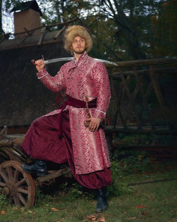 Photo for Confident brave young ukrainian cossack dressed ornate festive traditional clothing and fur hat holding saber on his shoulder looking away. Full length portrait - Royalty Free Image