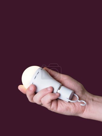 A switched-on LED rechargeable lamp in the hands of a person on a burgundy background