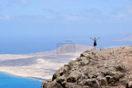 Foto de Mujer de pie con los brazos levantados en la cima de una colina. Con La Graciosa al fondo - Imagen libre de derechos