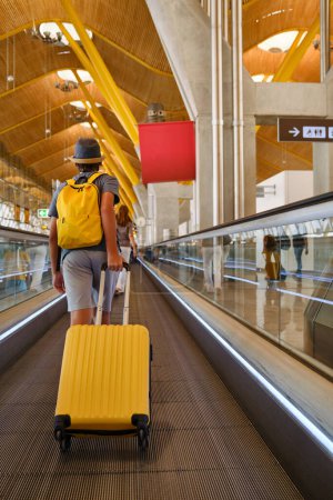 Foto de Full body back view of unrecognizable male tourist in hat with yellow suitcase and backpack walking on travolator in airport - Imagen libre de derechos