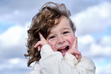 Retrato de chica alegre adorable con el pelo ondulado y sonrisa dentada mirando a la cámara mientras toca las mejillas con las manos cruzadas y con dos dedos mostrando signo V