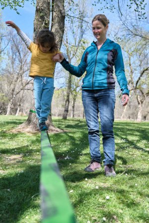 Smiling adult mother helping son balance on slackline tied to trunk and walk in park with green trees in daylight