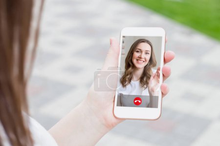 young caucasian woman holding a mobile phone in the street and talking to her girlfriend on video chat, video calls, conference. Caucasian girl waving her hand on the mobile phone screen