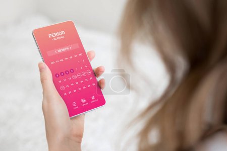 Téléchargez les photos : Femme tenant un téléphone portable avec une application de calendrier de cycle menstruel et assise dans sa chambre - en image libre de droit