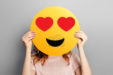Foto de Emoticon enamorado. Chica sostiene un sonriente amarillo con emoticono romántico cara de amor aislado sobre un fondo gris. Concepto de San Valentín - Imagen libre de derechos