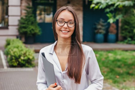 Photo for Student girl in glasses holds a folder and smiles against the background of the facade of the building. Remote education. Education abroad - Royalty Free Image