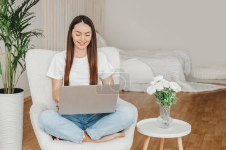 Photo for A young caucasian brunette woman sits at home on an armchair with a laptop and is trained in an online profession. Online school of English. Session with a psychologist - Royalty Free Image