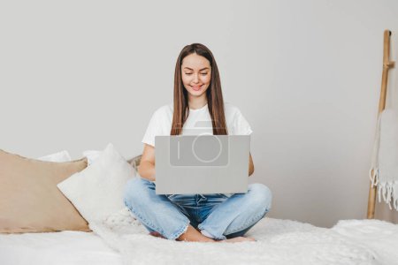 Chica comercializador de Internet se sienta en casa en la cama con un ordenador portátil y trabaja de forma remota en casa. Profesión independiente