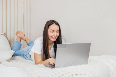 Téléchargez les photos : Une vendeuse travaille allongée sur un lit avec un ordinateur portable à la maison regardant l'écran et souriant. Étudiant prend des cours d'anglais en ligne - en image libre de droit