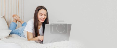 Téléchargez les photos : Une vendeuse travaille allongée sur un lit avec un ordinateur portable à la maison regardant l'écran et souriant. Étudiant prend des cours d'anglais en ligne - en image libre de droit