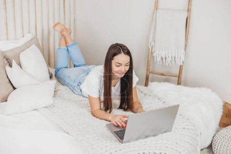 Téléchargez les photos : Gestionnaire de publicité fille travaille à la maison avec un ordinateur portable couché sur un lit blanc vue sur le dessus. Profession moderne à distance - en image libre de droit