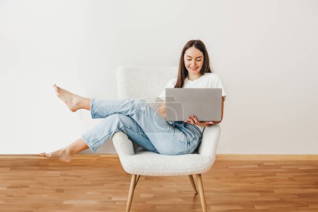 student girl using laptop at home. brunette woman sitting on chair smiling and look on computer, wear white shirt and jeans. Freelance concept