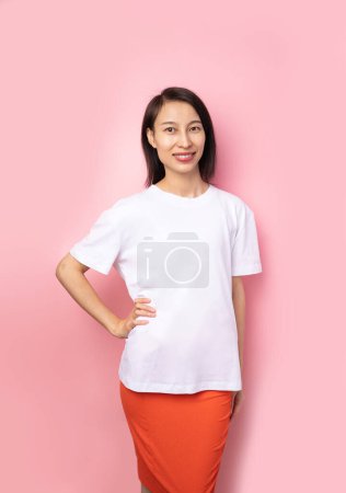 Photo for Beautiful smiling Asian woman in white blank solid color t-shirt looking at camera. Pink background. Empty copyspace template foe clothes. hand on waist - Royalty Free Image