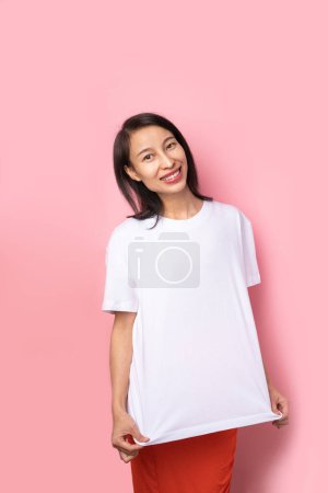 Photo for Adorable beautiful Asian woman in white blank solid color t-shirt looking at camera and smiling. Pink background. Empty copyspace template foe clothes - Royalty Free Image