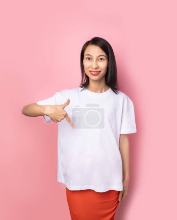 Photo for White t-shirt mockup. Adorable beautiful Asian woman in white blank solid color t-shirt looking at camera and smiling. Pink background. Empty copyspace template foe clothes - Royalty Free Image