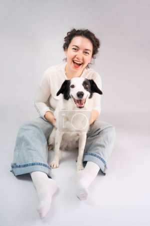 Photo for Happy laughing woman and her dog looking at camera sitting on grey floor. Owner young girl in blue jeans and white jumper having fun with her cute pet white young border collie - Royalty Free Image