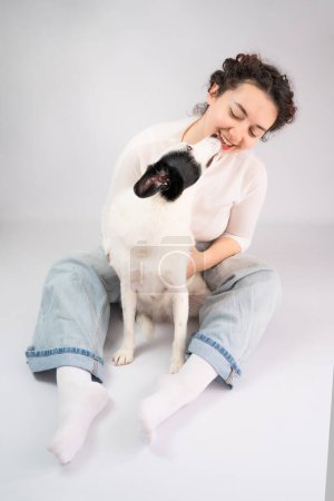 Photo for Pet kiss. Smiling young woman hugging her white dog sitting on the floor. Relaxed touching trusting relationship between dog and owner. Grey (gray) background. studio shot - Royalty Free Image