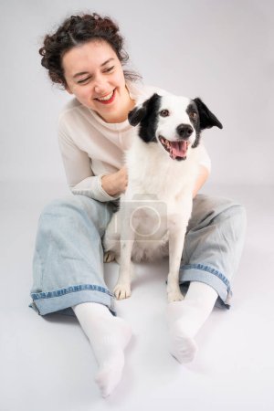 Photo for Dog and woman smiling sitting on gray background. Owner young girl in blue jeans and white jumper cuddling with her cute pet border collie young dog. Positive happy emotions. Wide open smile - Royalty Free Image