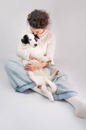 Photo for Dog owner and dog cuddling. Hugging young woman and adorable white border collie pet sitting on the floor. grey (gray) background. Casual blue jeans and white clothes. Best friends trust relationship - Royalty Free Image
