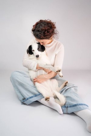 Photo for Woman hugging white dog sitting on grey background. Best friend moment of tenderness. Owner young girl in blue jeans and white jumper cuddling with her cute pet border collie young dog. Studio shot - Royalty Free Image