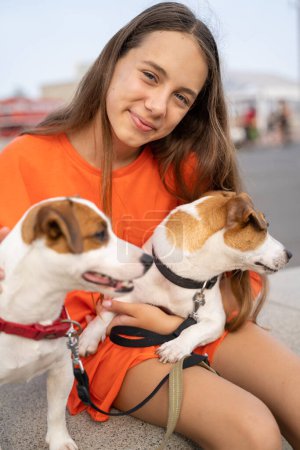 Foto de Hermosa adolescente mirando a la cámara abrazando acariciando a dos perros Jack Russell terriers acostados en sus vueltas. Retrato de amigos. Color naranja brillante camiseta ropa - Imagen libre de derechos