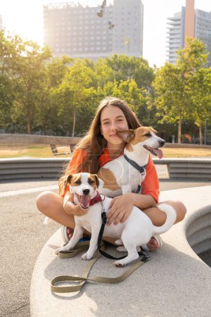 Foto de Chica adolescente en ropa de color naranja mirando a la cámara y sonriendo abrazando a sus dos adorables perros pequeños Jack Russell terrier blancos. Caminando en el parque urbano. sol de verano tarde - Imagen libre de derechos