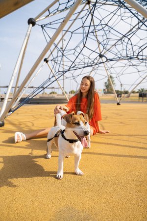 Foto de Paseo de verano con perro. Adolescente chica en naranja ropa sentado en el patio de recreo con su pequeño lindo blanco Jack Russell terrier chica en el fondo fuera de foco - Imagen libre de derechos