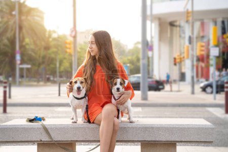 Foto de Chica con dos amigos perros. El ambiente de verano en la gran ciudad. Sonriente perfil de adolescente feliz en ropa naranja mirando lado abrazando mascotas lindas Jack Russells sentado en el banco. - Imagen libre de derechos
