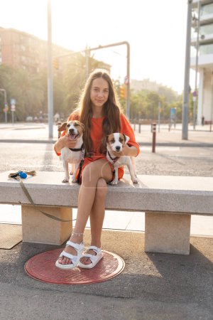 Foto de Hermosa chica adolescente de pelo largo sentada en el banco con sus dos perros terriers Jack Russell. Ropa deportiva casual de color naranja brillante. Tarde de verano paseando con perros en la gran ciudad. - Imagen libre de derechos