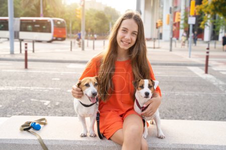 Foto de Chica con dos amigos perros. El ambiente de verano en la gran ciudad. Sonriente chica adolescente feliz en ropa naranja abrazando mascotas lindas Jack Russells sentado en el banco - Imagen libre de derechos
