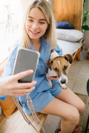 Foto de Chica guapa rubia tomando selfie con su pequeño perro Jack Russell terrier. Cómoda ropa azul con estilo. Almuerzo café en la cafetería. Influencer blogger live streaming. Fotografía de composición vertical. - Imagen libre de derechos