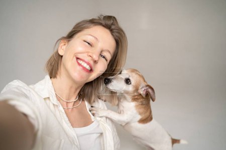 Foto de Adorables mejores amigos rubia mujer y perro pequeño Jack Russell terrier sonriendo y mirando a la cámara. divertirse la mascota. Un perro besador. camisa blanca y jeans azules - Imagen libre de derechos