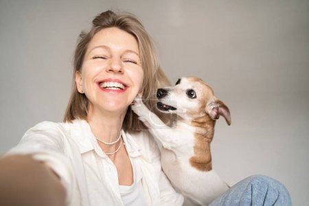 Foto de Selfie retrato de hermosa rubia sonriente feliz mujer y su pequeño perro lindo Jack Russell terrier mirando a la cámara y sonriendo. divertirse ingenio la mascota. ¡Los mejores amigos! camisa blanca y jeans azules - Imagen libre de derechos