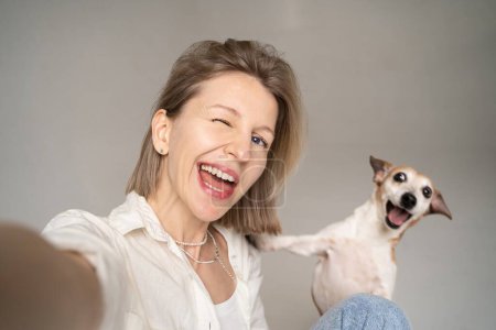 Foto de Feliz retrato selfie de la mujer rubia guiñando el ojo y su pequeño perro lindo Jack Russell terrier mirando a la cámara y sonriendo. divertirse con la mascota. ¡Los mejores amigos! camisa blanca y jeans azules - Imagen libre de derechos