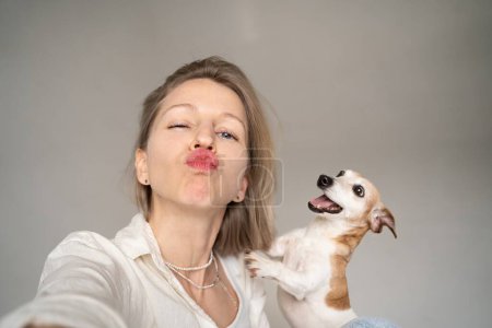 Foto de Besa los labios. Mujer rubia divertida y perro pequeño jugando a divertirse posando para retratos selfie, Adorable mascota pequeña mejor amigo. Estudio sobre fondo gris - Imagen libre de derechos
