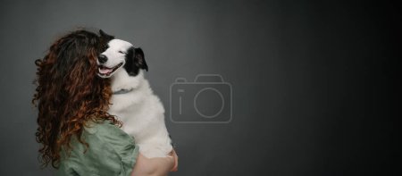 Adorable pet trust, love and support. Back of woman with long curly hair holding her white border collie dog happy smiling and looking at the camera Grey background studio shot. Long horizontal banner