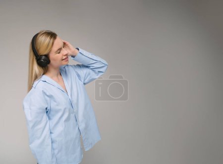 Mujer rubia sonriente usando auriculares perfil lateral escuchando música. Camisa azul de gran tamaño. Espacio de copia de fondo gris
