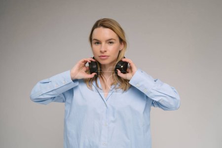 Young Caucasian blonde woman looks at the camera with serious concentration. holding headphones on her neck. Blue casual shirt. Grey background studio shot music audio theme