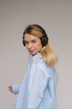 Listening to music. Young Caucasian blonde woman looks at the camera half a turn from  with flirting smile with headphones on her neck. Blue casual shirt. Grey background studio shot music audio theme