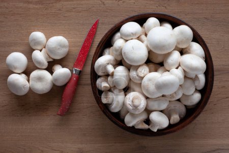 Photo for Fresh champignons on the table, in a bowl and on a wooden board with a red knife, top view of vegetable ingredients for cooking. - Royalty Free Image