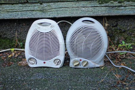 deux ventilateurs de chauffage électrique en plastique blanc gris se tiennent sur l'asphalte près d'un mur en bois vert dans la rue