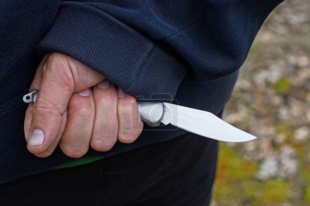 hand of a male criminal in blue clothes and a gray knife behind his back on the street