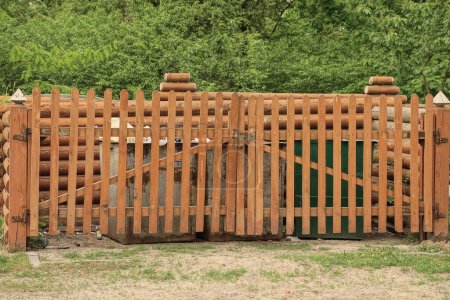 Foto de Parte de una cerca de madera marrón con una puerta cerrada hecha de tablas frente a los botes de basura en la calle contra el telón de fondo de la vegetación verde - Imagen libre de derechos