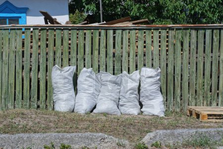 Téléchargez les photos : Une rangée de grands sacs en plastique blanc cinq avec des ordures près d'un mur de clôture en bois vert dans l'herbe sur une rue rurale - en image libre de droit
