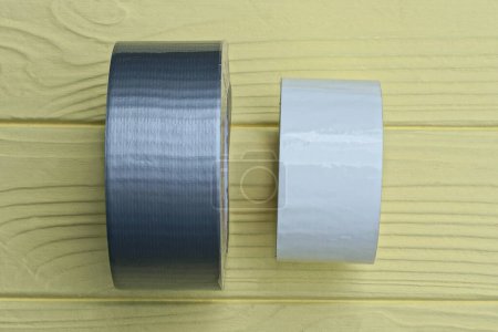 Foto de Un rollo de cinta gris reforzada y un rollo de cinta blanca yacen sobre una mesa de madera amarilla - Imagen libre de derechos