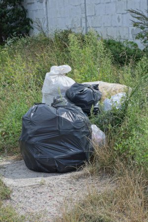 Foto de Una pila de basura de bolsas de plástico completas blancas y negras en tierra gris entre hierba verde afuera - Imagen libre de derechos