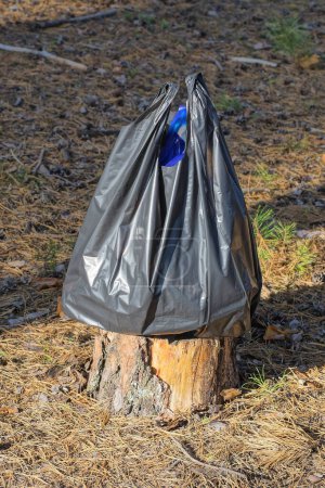 Téléchargez les photos : Un grand sac en plastique noir déchiqueté avec des ordures debout sur une souche grise à l'extérieur dans la nature - en image libre de droit