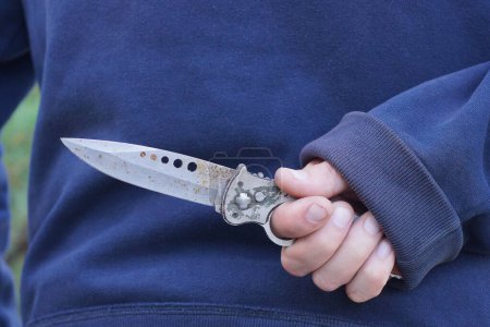 Foto de Mano de un criminal con ropa azul y un cuchillo gris oxidado detrás de su espalda en la calle - Imagen libre de derechos